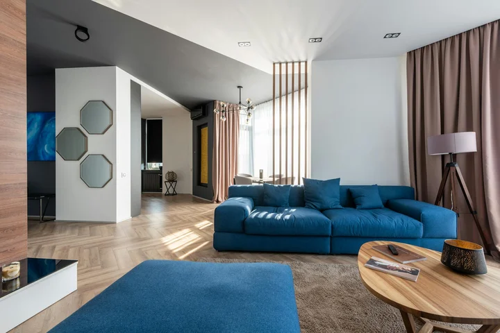 Modern living room with a blue sectional sofa, light wood flooring, a round wooden coffee table, and floor-to-ceiling curtains bathed in natural light.