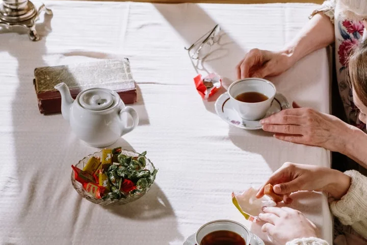 Hands of two people at a white-table tea setup with a teapot, cups of tea, and a lemon slice on the table