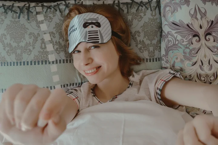 Woman wearing a sleep mask lying in bed, reaching toward the camera
