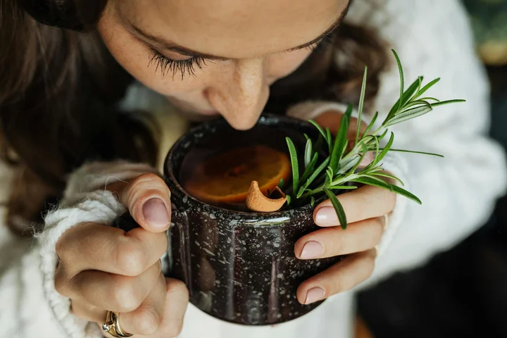 Close-up of a person smelling a mug of hot beverage, hands cupping a dark mug with sprigs of rosemary.
