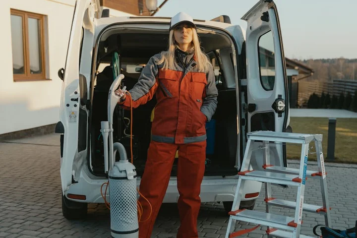 A field technician wearing orange and gray safety gear stands beside an open van filled with cleaning equipment, including a large cylinder and a step ladder, illustrating modern concrete cleaning methods.
