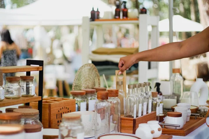 Outdoor market stall with glass jars and cleaning bottles on a wooden table; a hand reaches toward an item.