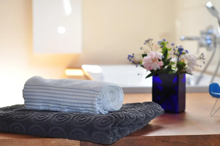 A rolled white towel sits atop a dark patterned towel on a wooden bathroom counter, with a blue vase of flowers and a blurred sink in the background.