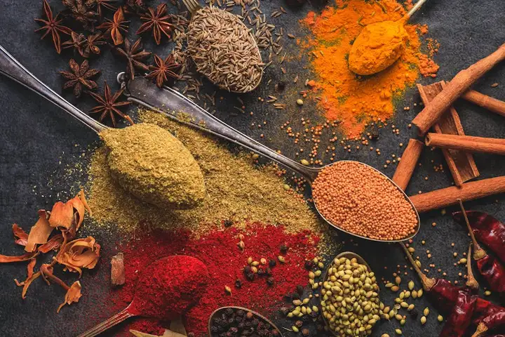 Assorted ground spices on a dark surface, featuring bright yellow turmeric powder, red chili powder, curry powder, cinnamon sticks, star anise, and seeds.