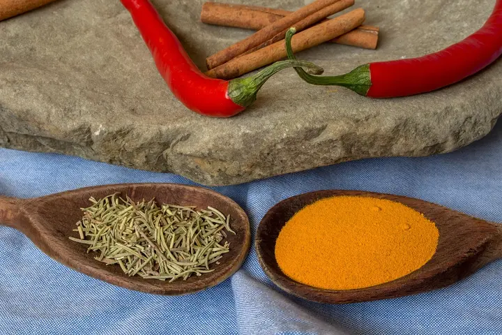 Red chili peppers and cinnamon sticks on a rock surface, with two wooden spoons on a blue cloth; one spoon contains dried rosemary and the other holds turmeric powder