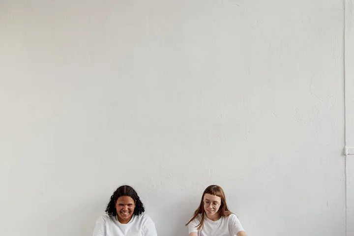 Two people sit on the floor against a plain light wall, smiling.