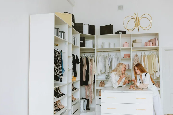 Two women stand at a white island in a bright walk-in closet, sorting and discussing clothing as shelves filled with outfits and accessories surround them.
