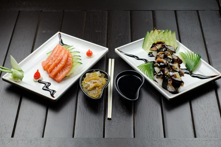 Two white plates with sashimi and cooked fish on a dark wooden table, with chopsticks and dipping sauces nearby.