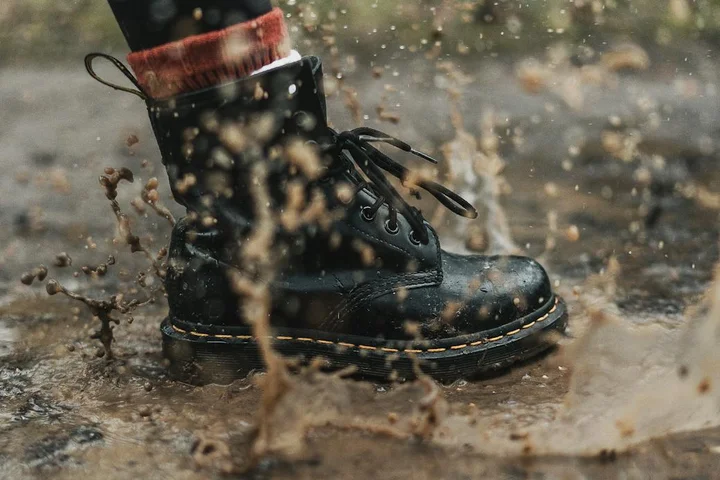 Close-up of a black UGG-style boot with a brown fur trim stepping through muddy water, with splashes around.
