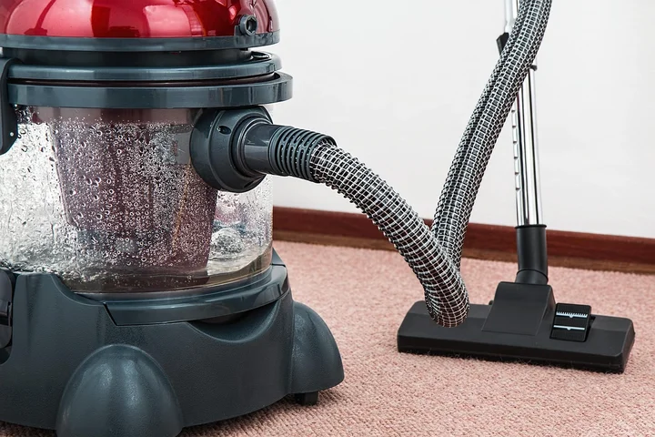 A vacuum cleaner with a clear dust canister and a flexible hose on a pink carpet, ready to remove glitter.