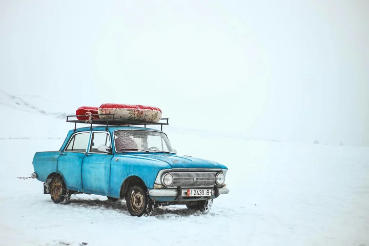 Blue vintage car in a snowy landscape with a roof rack and luggage on top, showing weathered paint.