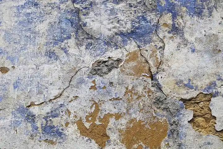 A weathered interior wall with peeling paint and damaged plaster, showing layers of blue, white, and tan.