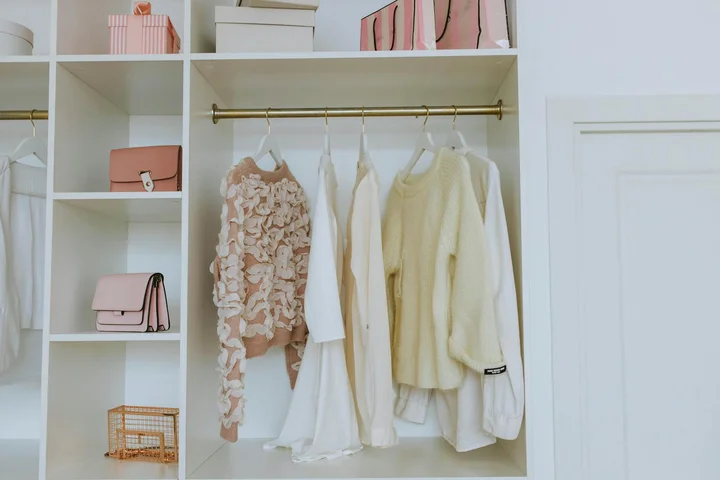 Open wardrobe with white shelving, hanging light-colored garments, and shelves displaying bags and boxes.