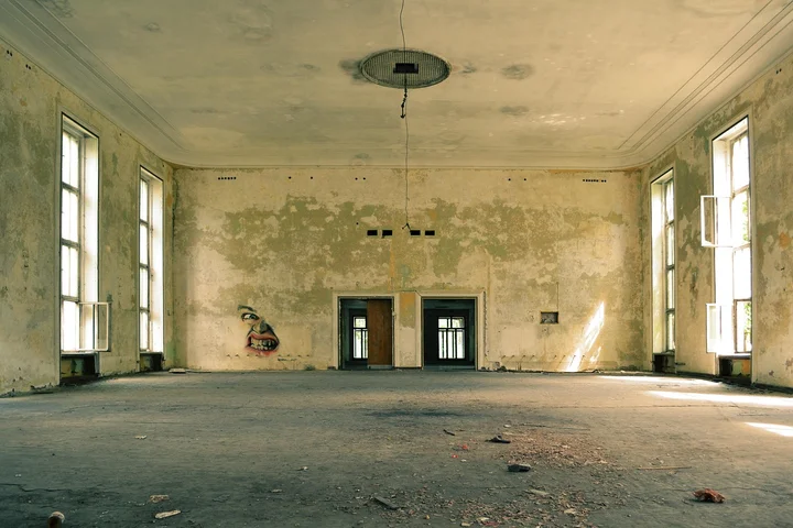 Room with peeling plaster and water stains on the ceiling and walls, large windows along the sides