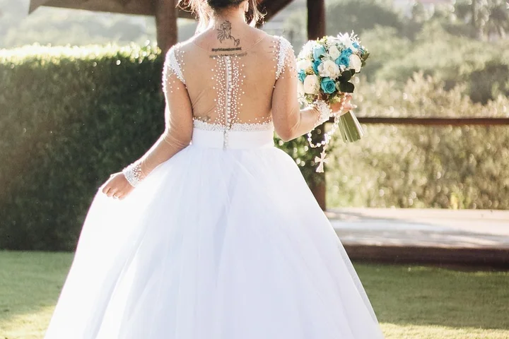 Back view of a bride wearing a white wedding gown with intricate beadwork along the back, outdoors with a bouquet.