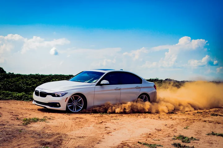 White sedan driving on a dusty dirt road, kicking up a cloud of brown dust under a blue sky.
