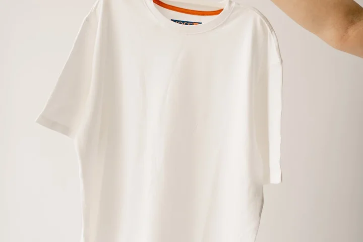 A plain white T-shirt being held up against a neutral background, showing clean fabric.