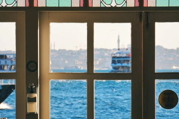 View through a glass window/door of blue ocean water with boats in the distance, sunlight streaming in