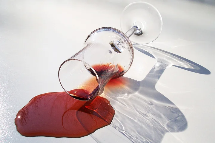 A wine glass lies on its side on a white surface, spilling red wine that forms a stain and casts a shadow.