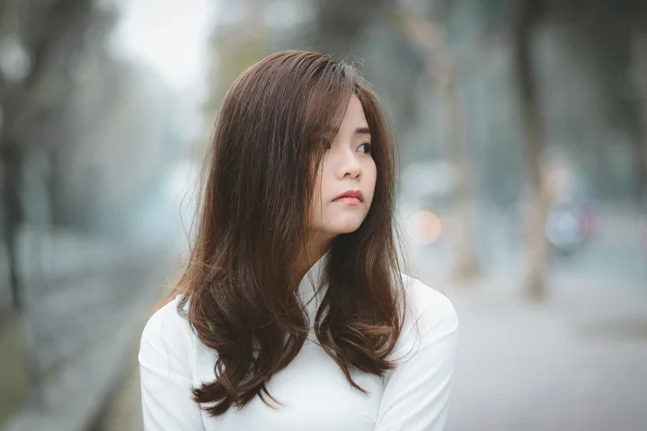Portrait of a woman with long brown hair wearing a white top, standing outdoors in an urban setting, looking to the side.