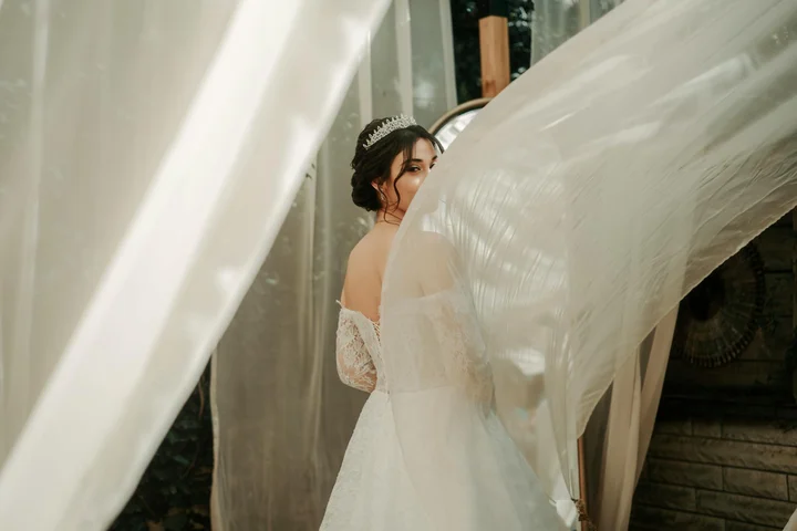 A woman in a wedding dress stands behind sheer, flowing drapes indoors, with fabric billowing around her.